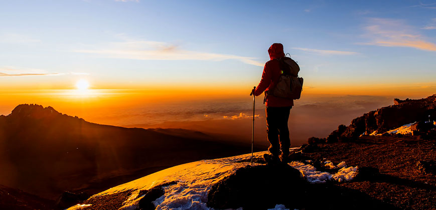 Man with backpack and trekking poles celebrates summit of Mount Kilimanjaro at sunrise, gazing over Mount Meru – high-altitude adventure with Pizet Safaris